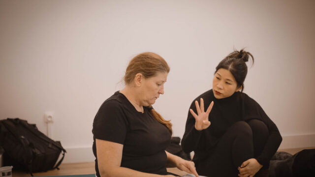 Lara, professeur de Hatha Yoga, Yin Yoga et Yin Yang Yoga, explique verbalement une posture à une élève qui tient ses notes lors d'une session de formation 200h à l'Académie Yoga Paris.