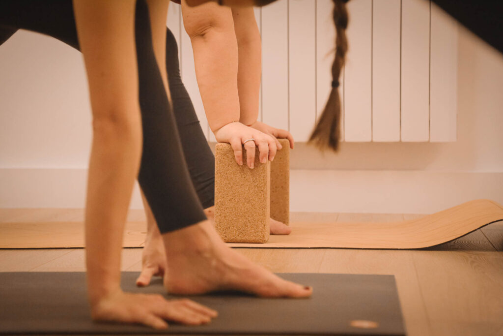 Lors d'un cours de Yoga une main posée sur une brique de yoga en liège sur un tapis Manduka. Au premier plan flou, une autre personne a la main posée au sol lors d'une formation de Yoga à Paris 15.