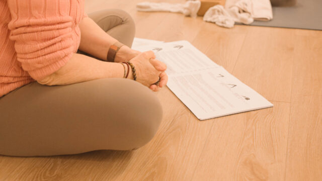 Plan rapproché d’une personne assise, les mains posées sur les genoux à côté du manuel des postures de yoga pendant la formation 200h Hatha Yoga au studio du XVe arrondissement de Paris.